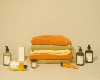 Three folded towels in orange, green, and yellow sit center stage on a beige background, surrounded by skincare bottles—including FRAMA Body Wash, 375ml—along with various jars and tubes featuring minimal labels.