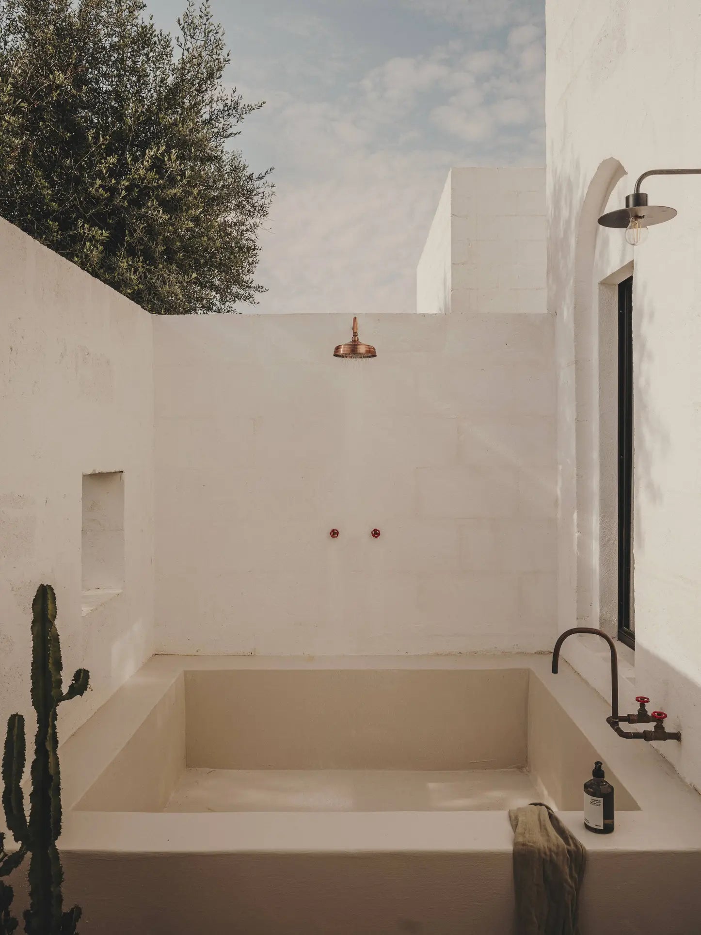Minimalist outdoor bathtub set by white stucco walls, featuring a copper shower head and faucet, a small cactus, a towel, and FRAMA Body Wash (375ml) from FRAMA—bathed in natural sunlight under a partly cloudy sky.