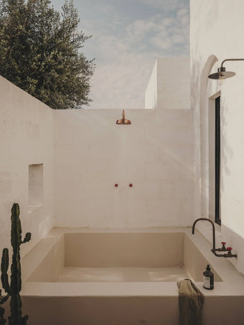 Minimalist outdoor bathtub set by white stucco walls, featuring a copper shower head and faucet, a small cactus, a towel, and FRAMA Body Wash (375ml) from FRAMA—bathed in natural sunlight under a partly cloudy sky.