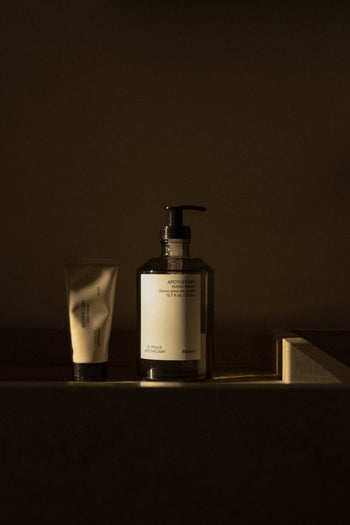 Two FRAMA skincare products—a tube and a 375ml Hand Wash pump—rest side by side in dim, moody lighting. The partially visible labels suggest organic ingredients and sandalwood scent but aren’t fully readable.