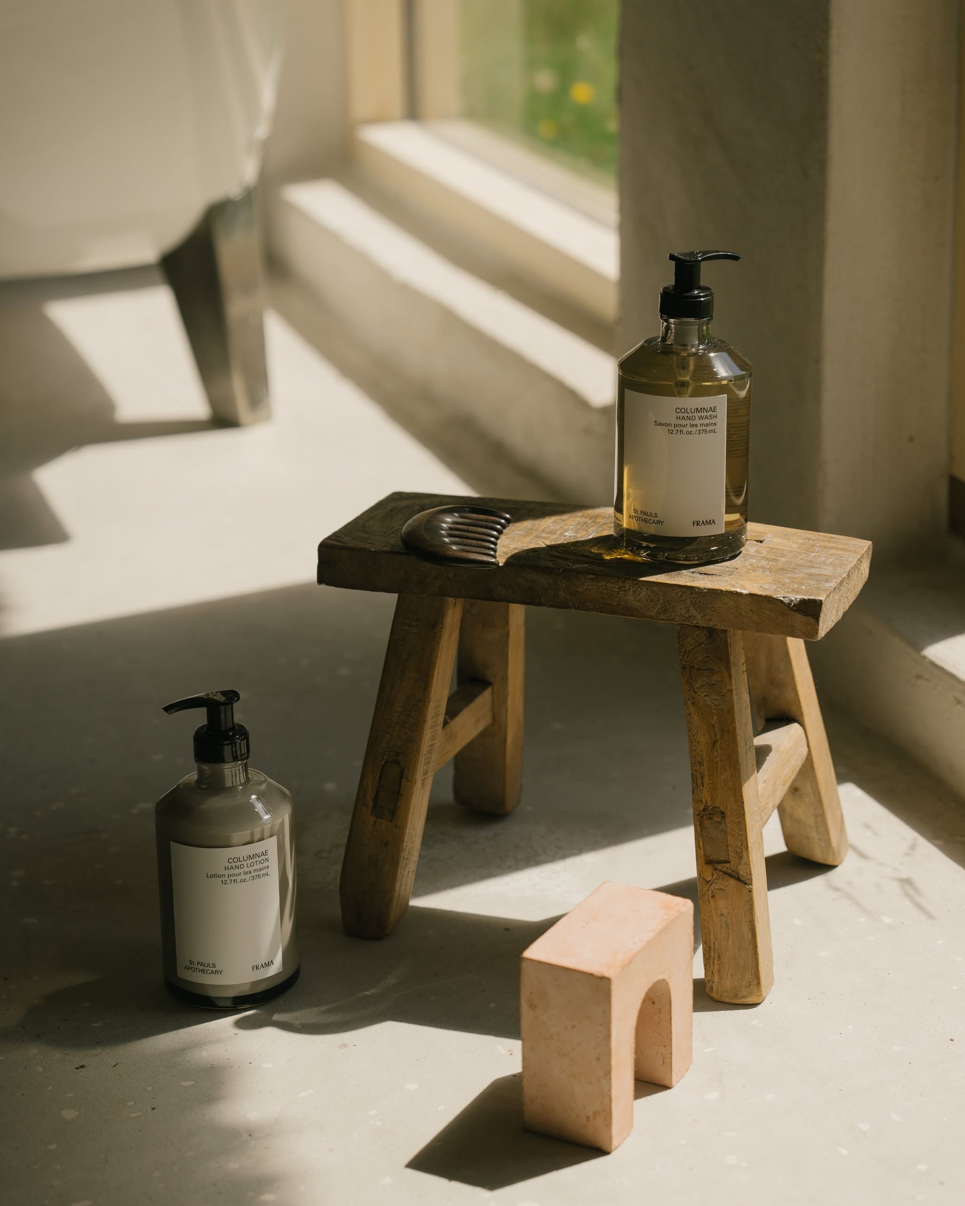 A wooden stool by a sunlit window holds a brown-pump FRAMA Hand Wash, 375ml, and a dark comb; another bottle and a small pink arch-shaped object sit on the floor beside it.