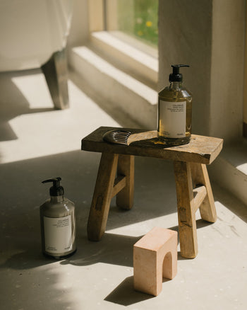 A wooden stool by a sunlit window holds a brown-pump FRAMA Hand Wash, 375ml, and a dark comb; another bottle and a small pink arch-shaped object sit on the floor beside it.