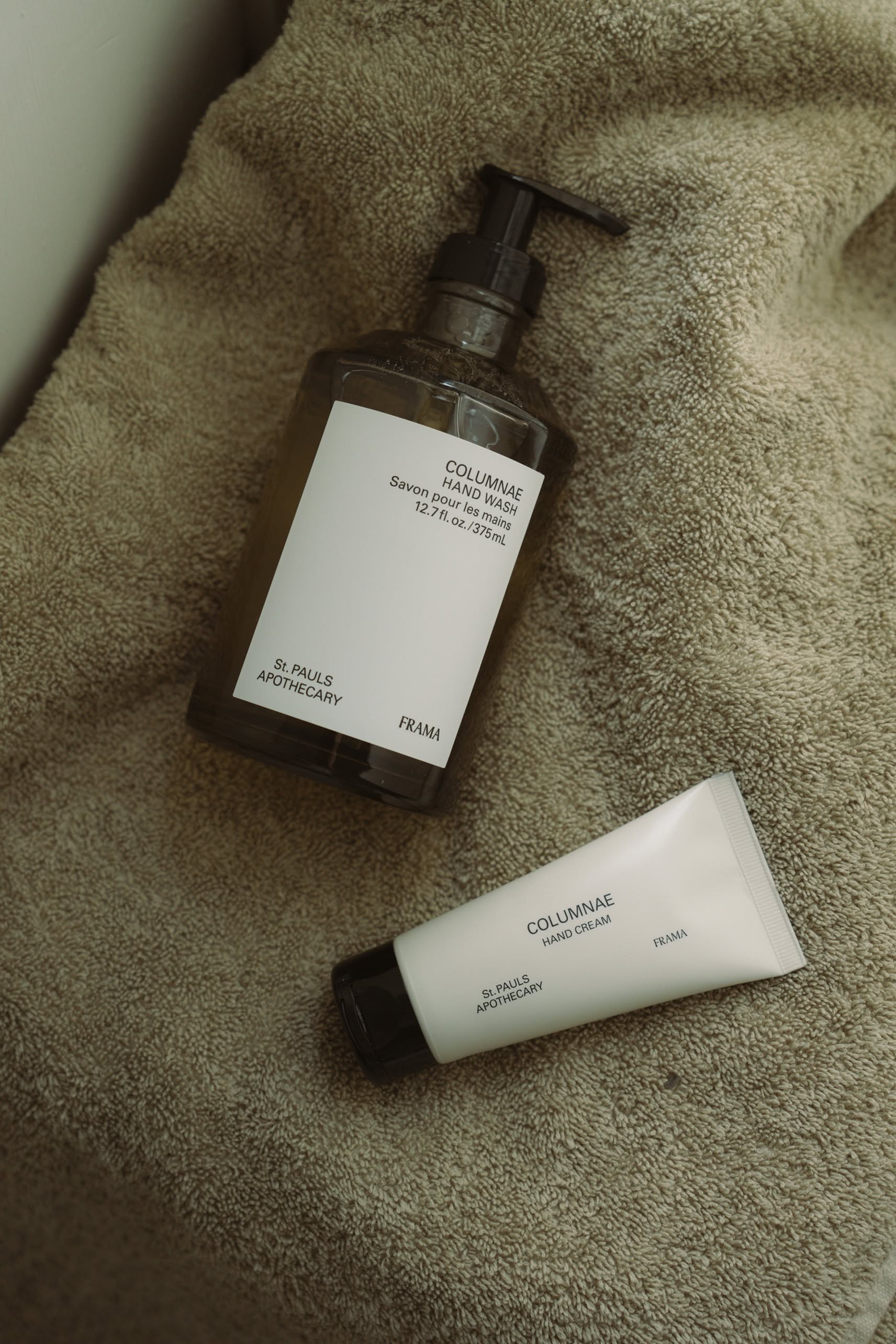 A bottle of FRAMA Hand Wash, 375ml, and a matching hand cream tube from FRAMA sit atop a soft beige towel.