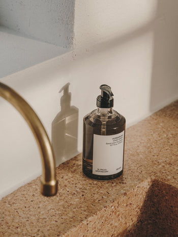 A 375ml FRAMA Hand Wash bottle with pump dispenser rests on a terrazzo countertop beside a brass faucet, casting a shadow on a white wall in gentle natural light.