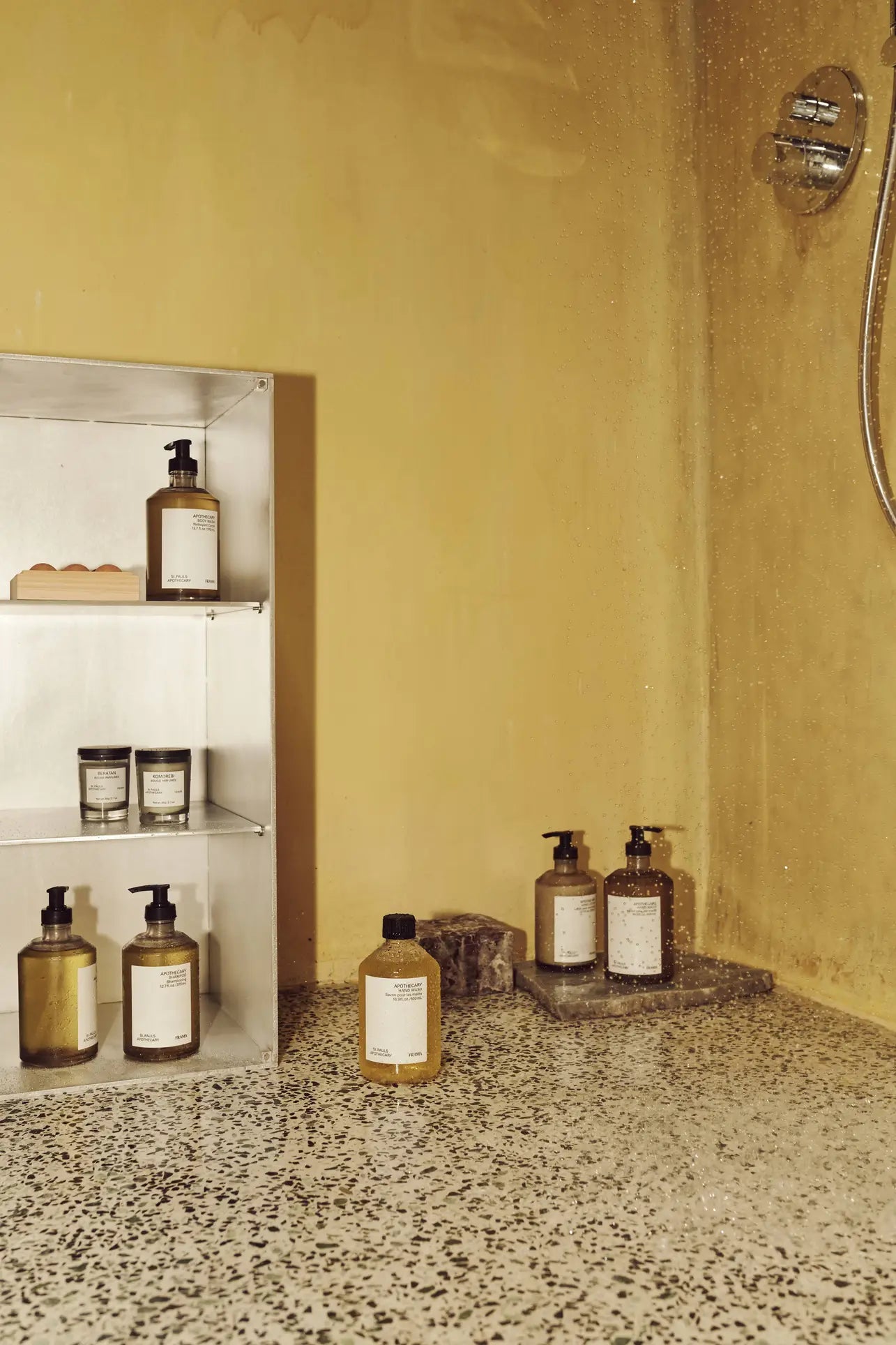 A shower area with yellow walls features a metal shelf holding various bottles, including FRAMA Hand Wash (500ml), and Hello Kitty wooden collectibles displayed on the terrazzo floor near a wall-mounted showerhead.