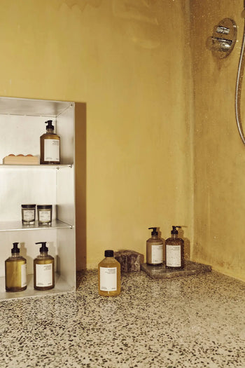 A shower area with yellow walls features a metal shelf holding various bottles, including FRAMA Hand Wash (500ml), and Hello Kitty wooden collectibles displayed on the terrazzo floor near a wall-mounted showerhead.