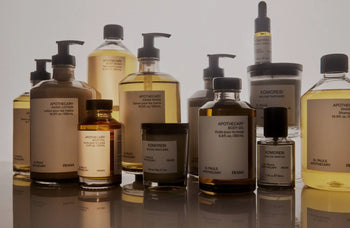 A 500ml FRAMA Hand Wash in an amber glass bottle with a minimalist white label is arranged on a neutral background beside wooden collectibles.