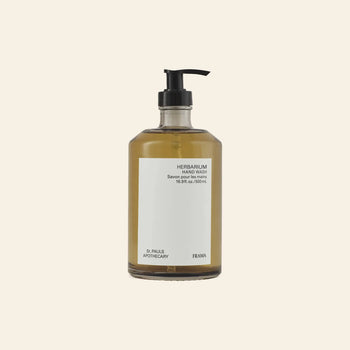 A clear pump bottle of FRAMA Hand Wash, 500ml, sits beside charming wooden collectibles against a plain beige background. Amber-colored liquid enhances its elegant look, and the minimalist label highlights the FRAMA brand.