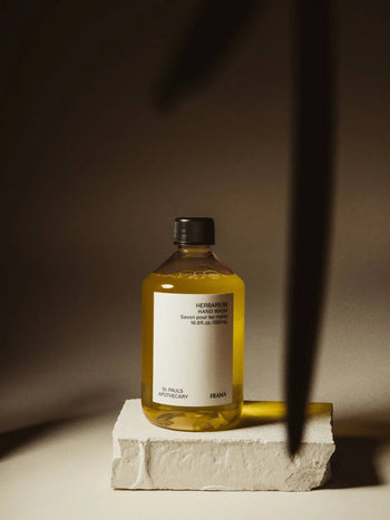 A clear bottle of FRAMA Hand Wash, 500ml with a black cap rests on a textured white block in warm, moody light. Shadows and wooden accents add charm to the scene as the FRAMA branding stands out against the partially covered background.