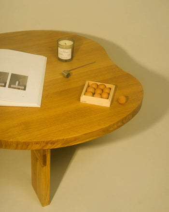 A wooden table displays an open book, the FRAMA 1917 Scented Candle (170g) glowing warmly, a small metal tool, and a box of round wooden balls, evoking Mediterranean charm on a light surface.
