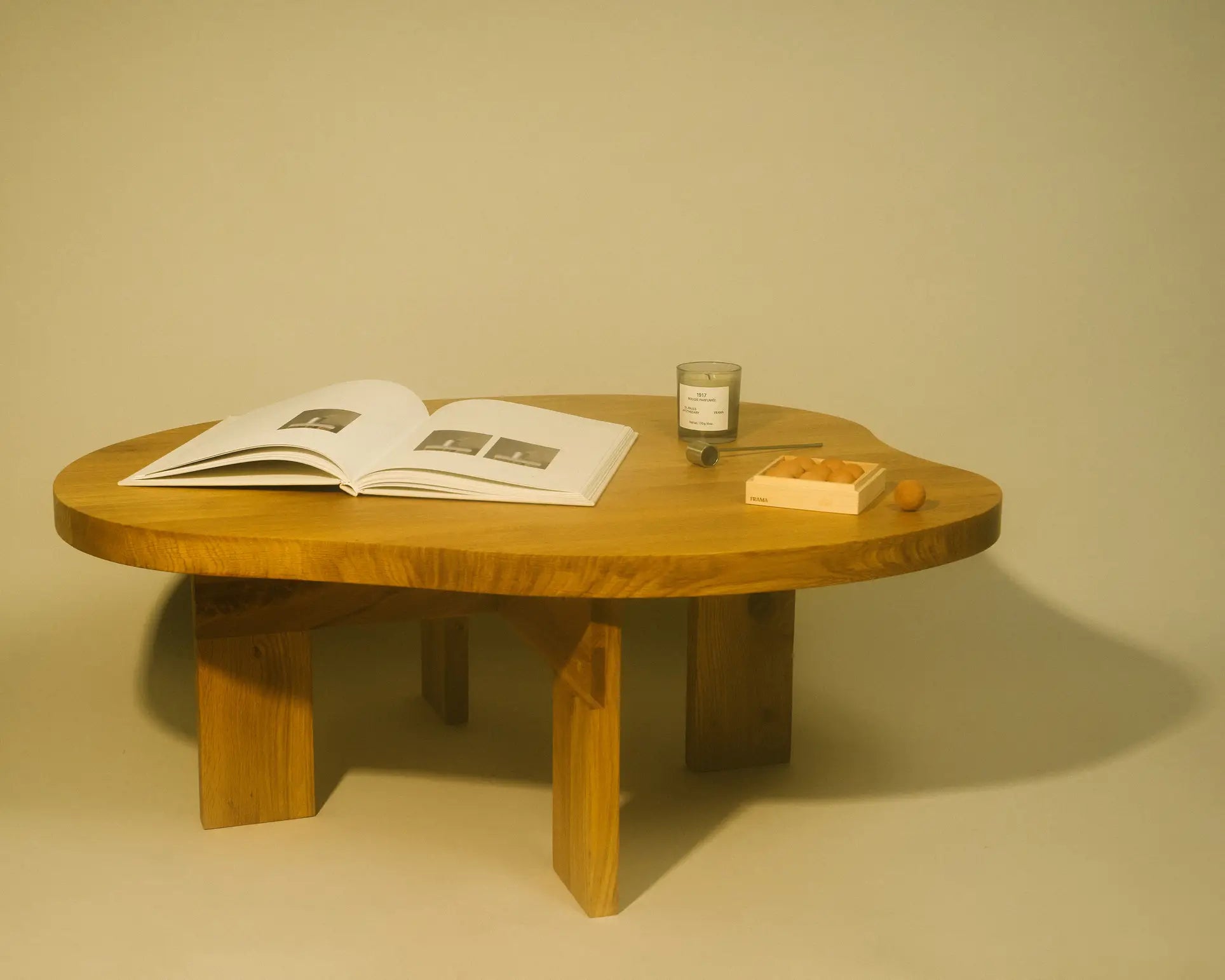 A wooden coffee table displays an open book, a FRAMA 1917 Scented Candle (170g), a small tray, and three apricots, all set against a plain beige background. The candle evokes classic Chypre and Mediterranean flora.
