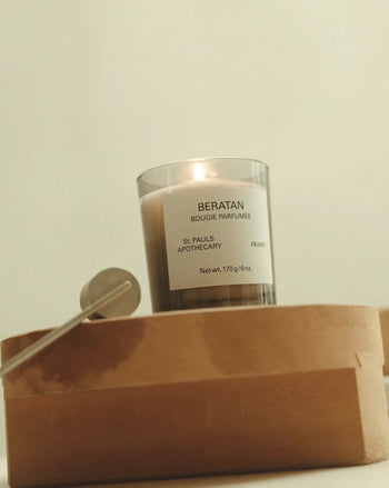 A FRAMA Beratan Scented Candle, 170g, labeled BERATAN BOUGIE PARFUMÉE rests on a wooden box beside a silver snuffer, filling the space with a warm tropical fragrance against a plain background.