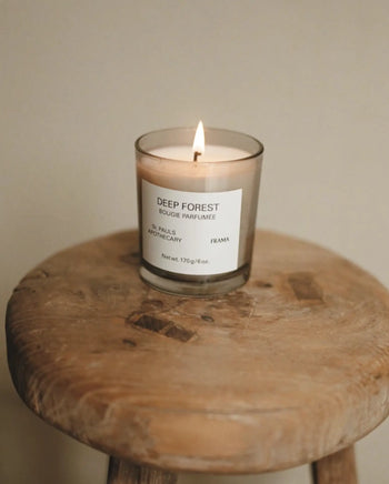 A lit FRAMA Deep Forest Scented Candle, 170g, in a glass jar rests on a rustic wooden stool, filling the air with an earthy Abies fir woods-inspired aroma against a neutral background.