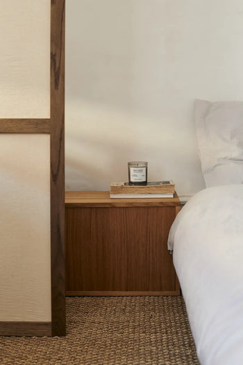 A cozy bedroom scene with a wooden nightstand displaying the FRAMA Deep Forest Scented Candle, 170g—by FRAMA—surrounding the space with an earthy Abies fir woods aroma, plus two books by a white bed and pillow on a woven carpet.