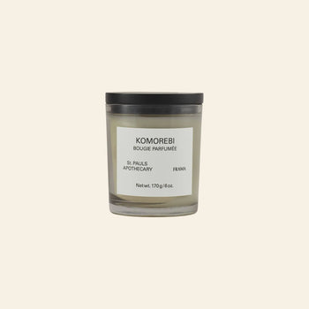 The FRAMA Komorebi Scented Candle, 170g, comes in a glass jar with a black lid and label, featuring an evocative aroma. Displayed against a plain beige background.