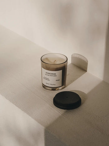 The FRAMA Komorebi Scented Candle, 170g, sits on a white surface in soft natural light. Its black lid casts a subtle shadow, creating a minimal and calm scene that hints at its gentle floral aroma.