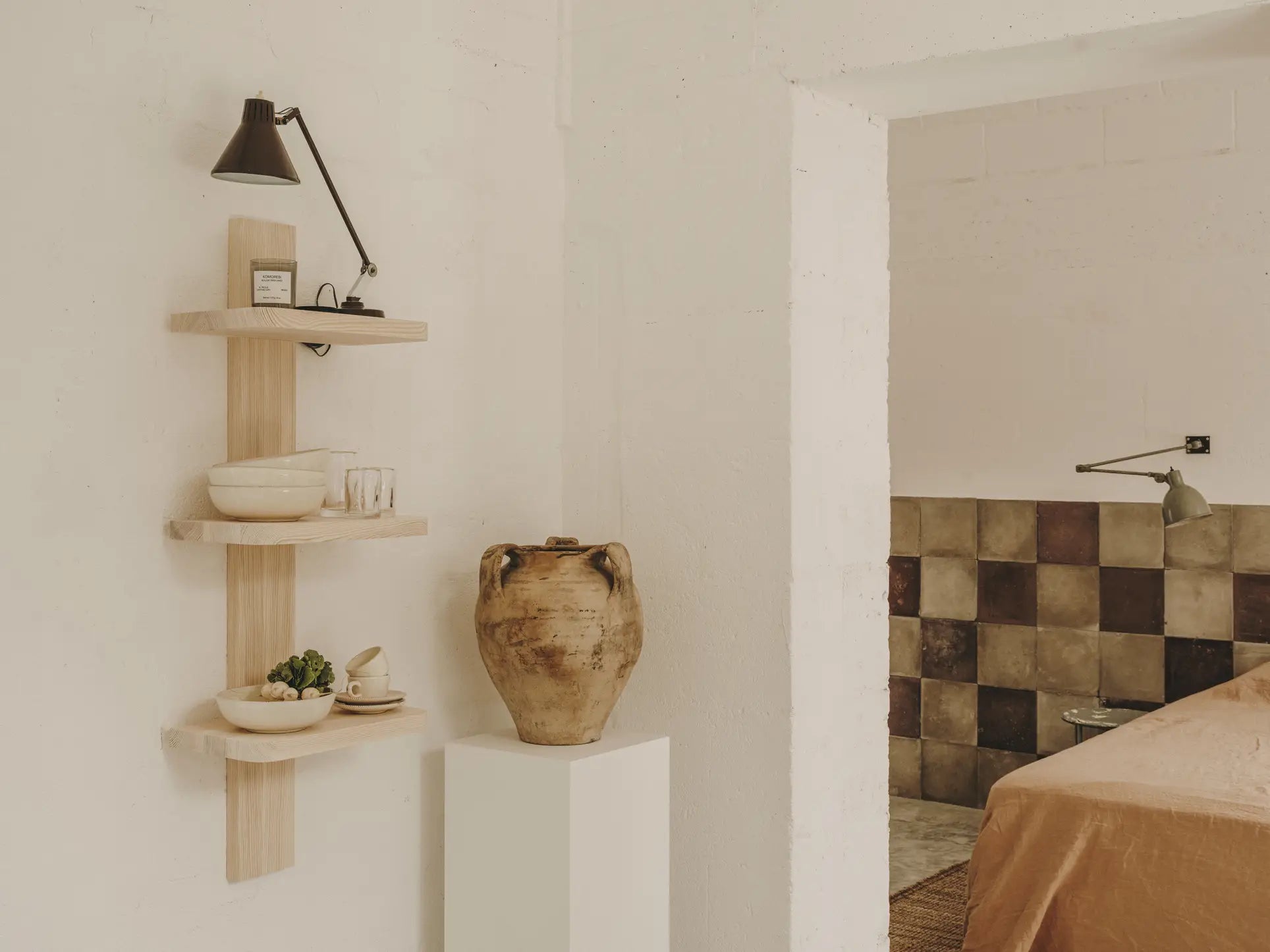 A minimalist interior with light wooden shelves displays ceramics, a succulent, and glasses. The space is filled with the evocative aroma of the FRAMA Komorebi Scented Candle, 170g. Brown and beige tiles and a tan bedspread appear in the next room.