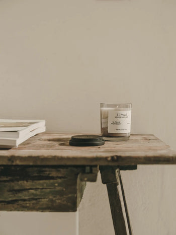 A rustic wooden table displays a closed black jar, stacked white papers or magazines, and a lit FRAMA St. Pauls Scented Candle (170g) in a glass container against a plain off-white wall.