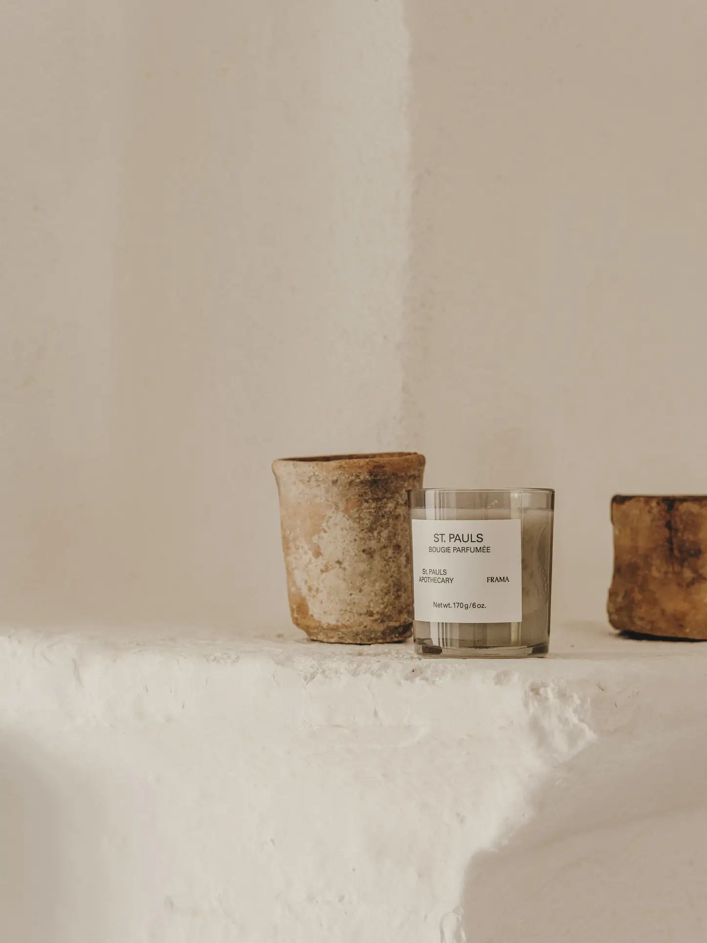 A minimalist scene featuring the FRAMA St. Pauls Scented Candle, 170g, displayed between two rustic clay pots on a textured white ledge against a beige wall, capturing the timeless charm of FRAMA’s Apothecary Collection.