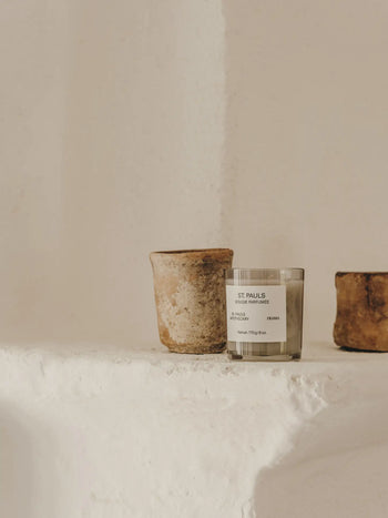 A minimalist scene featuring the FRAMA St. Pauls Scented Candle, 170g, displayed between two rustic clay pots on a textured white ledge against a beige wall, capturing the timeless charm of FRAMA’s Apothecary Collection.