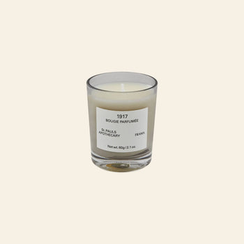 The FRAMA 1917 Scented Candle, 60g, features white wax in a small glass vessel and evokes Mediterranean flora and chypre notes, shown against a plain light beige background.