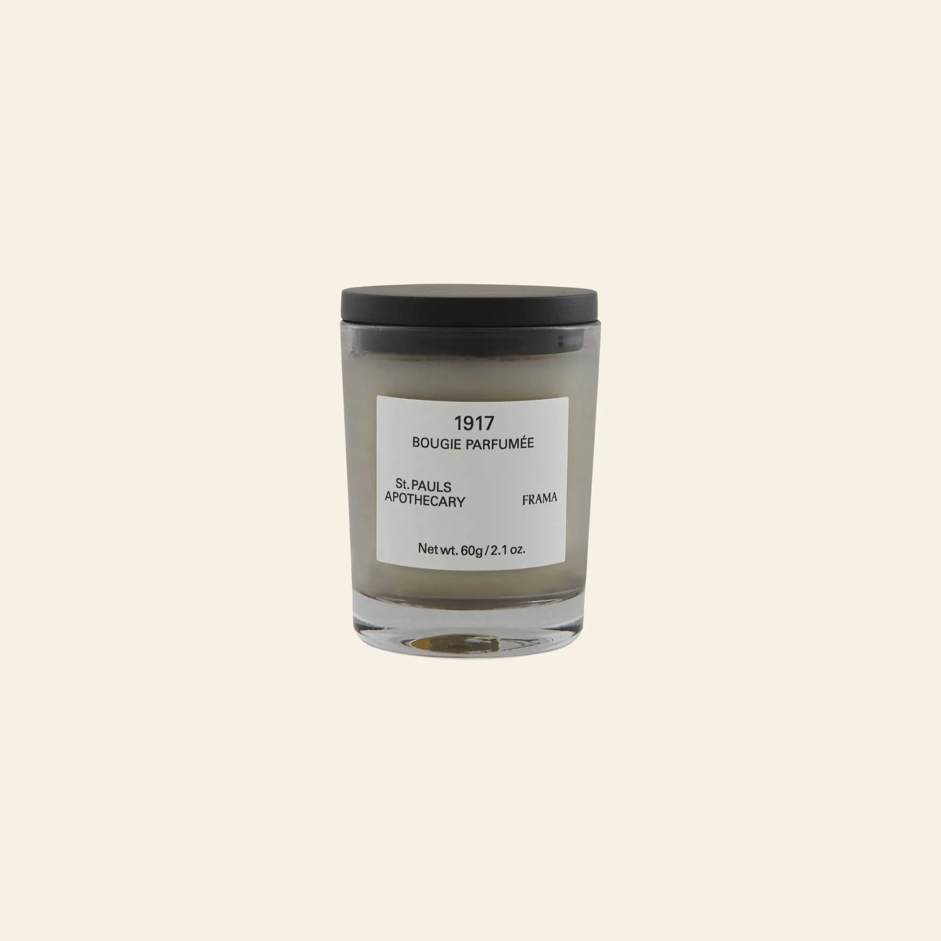 A small FRAMA 1917 Scented Candle (60g) in a glass jar with a black lid and minimalist label is displayed on a plain beige background, inspired by Mediterranean flora.
