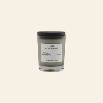 A small FRAMA 1917 Scented Candle (60g) in a glass jar with a black lid and minimalist label is displayed on a plain beige background, inspired by Mediterranean flora.