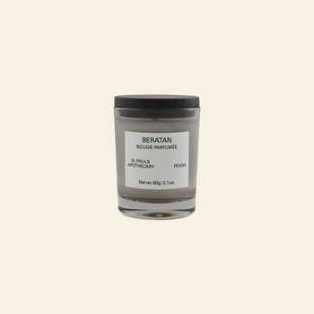 A minimalist white FRAMA Beratan Scented Candle, 60g, in a glass jar with black lid and label, offers a warm botanical fragrance. The candle is set against a plain beige background.