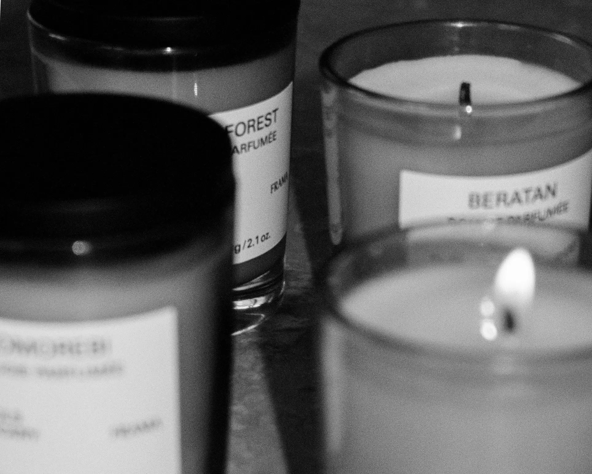 Four FRAMA candles in glass jars, including a FRAMA Beratan Scented Candle (60g), are arranged closely; one candle burns, filling the scene with warm fragrance. The black and white image shows blurred, partially visible candle labels.