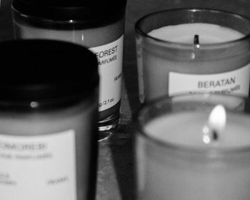 Four FRAMA candles in glass jars, including a FRAMA Beratan Scented Candle (60g), are arranged closely; one candle burns, filling the scene with warm fragrance. The black and white image shows blurred, partially visible candle labels.