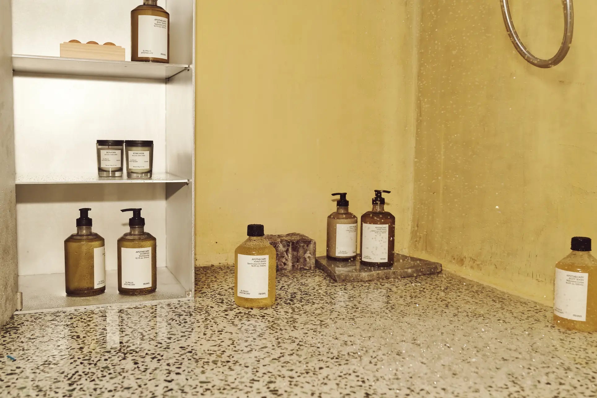 A minimalist bathroom with terrazzo flooring, a metal shelf for bath essentials, and soap dispensers on a stone tray against a yellow wall is enhanced by the warm fragrance of the FRAMA Beratan Scented Candle, 60g by FRAMA.