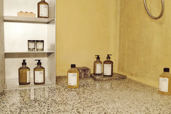 A minimalist bathroom with terrazzo flooring, a metal shelf for bath essentials, and soap dispensers on a stone tray against a yellow wall is enhanced by the warm fragrance of the FRAMA Beratan Scented Candle, 60g by FRAMA.