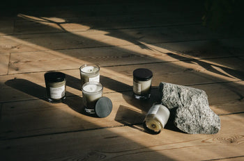 Five small glass candles, including the FRAMA Deep Forest Scented Candle 60g with an earthy Abies fir scents, are arranged on a wooden floor beside a large gray stone. Sunlight creates warm, natural shadows across the scene.