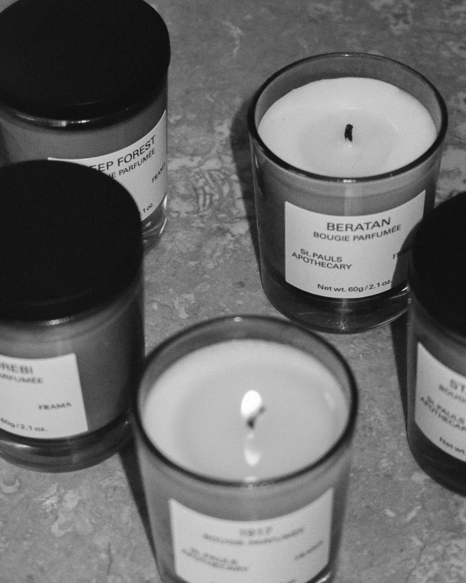 Black and white photo of five FRAMA candles with minimalist labels, including the FRAMA Deep Forest Scented Candle, 60g. Arranged on a textured surface, some are lit while others remain unlit, evoking earthy notes inspired by Abies fir woods.