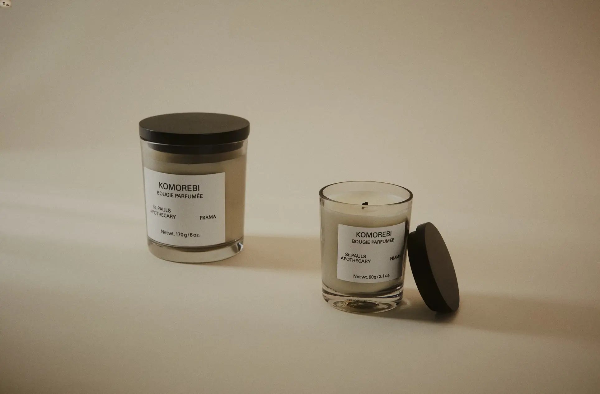 Two FRAMA Komorebi Scented Candles, 60g each, in minimalist glass—one open with its lid nearby, the other closed—rest against a plain beige background, suggesting a subtle floral aroma.