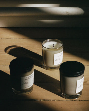 Three FRAMA Komorebi Scented Candles, 60g—two with black lids and one open—are arranged on a wooden floor bathed in sunlight and shadows, evoking warmth and a subtle floral scent.