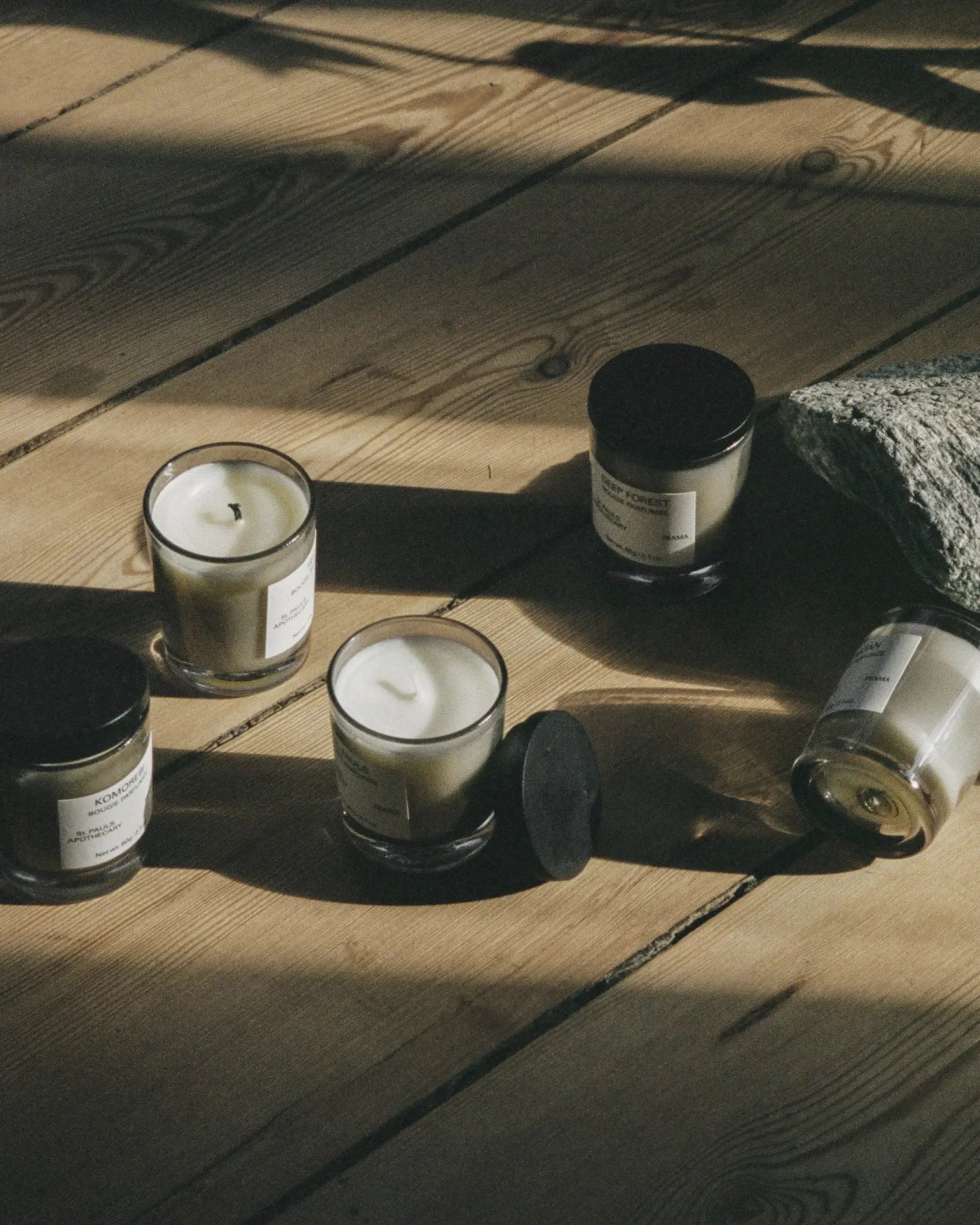 Five FRAMA St. Pauls Scented Candles, 60g each, with white wax and black lids—some open, some closed—rest on a sunlit wooden floor beside textured stone, their aroma mingling with the play of light and shadow.