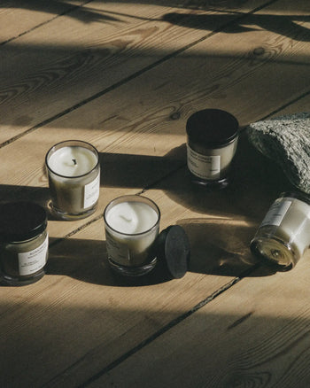 Five FRAMA St. Pauls Scented Candles, 60g each, with white wax and black lids—some open, some closed—rest on a sunlit wooden floor beside textured stone, their aroma mingling with the play of light and shadow.