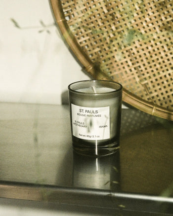 A FRAMA St. Pauls Scented Candle, 60g sits on a reflective surface beside a woven rattan object. Soft green leaves and gentle light capture the calm, minimalistic ambiance of FRAMA’s Apothecary Collection.