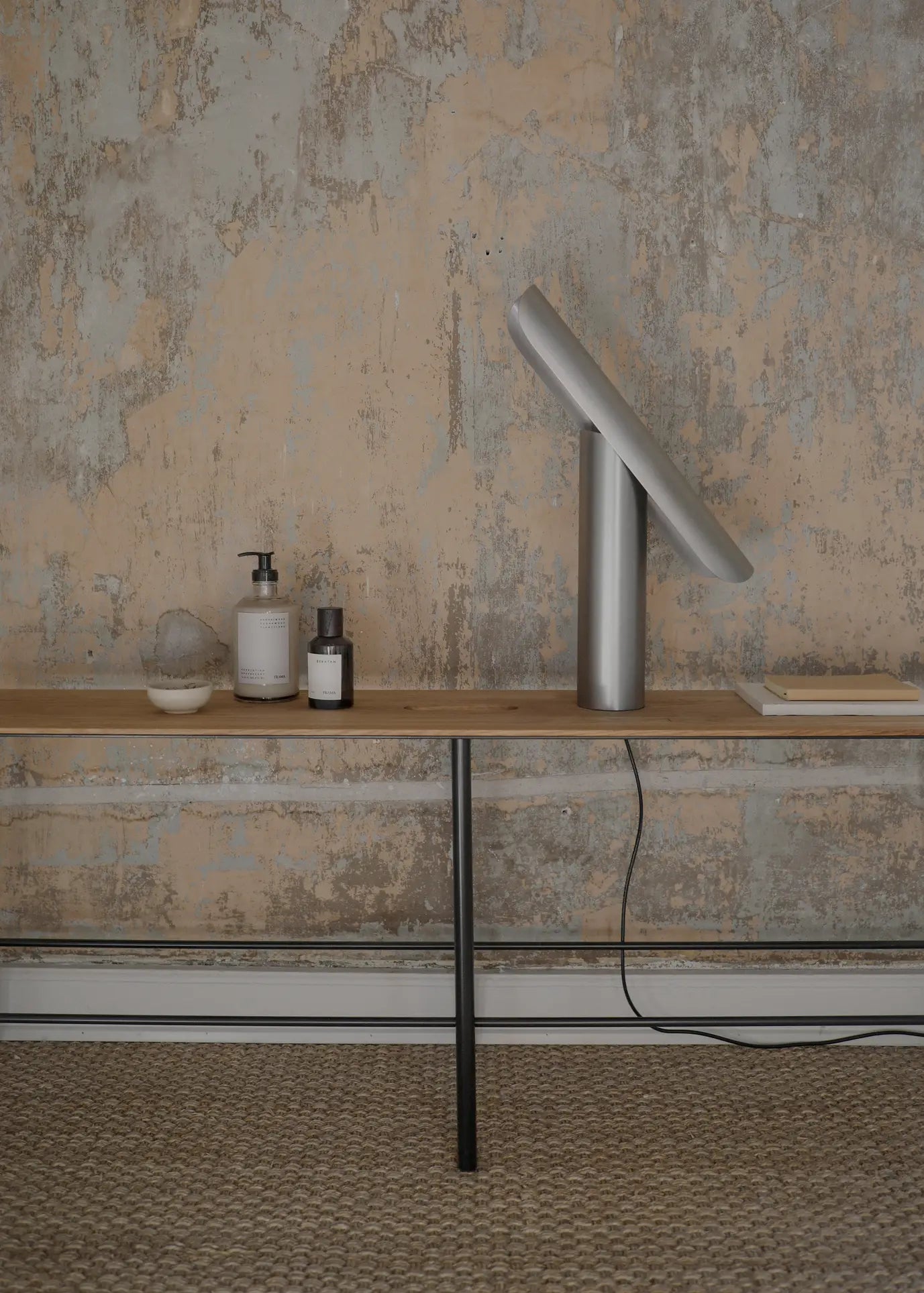 A minimalist wooden table stands against a textured wall. On it sits the FRAMA T-Lamp Table Lamp by FRAMA, featuring a slanted metallic shade and tilt mechanism, along with bottles, a closed book, and a hanging cord.