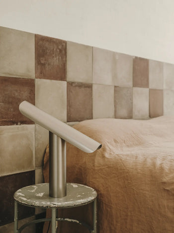 A minimalist bedroom features a beige and brown checkered tile headboard, a bed with a tan blanket, and the FRAMA T-Lamp Table Lamp by FRAMA on a round, weathered side table.