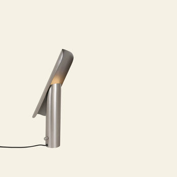 The FRAMA T-Lamp Table Lamp by FRAMA features a modern minimalist geometric design with a curved metallic shade and cylindrical base, emitting warm light. This contemporary desk lamp is plugged in and displayed against a light beige background.