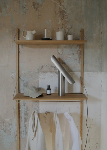 A minimalist wooden shelf unit against a textured wall holds the FRAMA T-Lamp Table Lamp, a hat, a small bottle, and hanging white garments. The top shelf displays a pitcher, candle, and cups with the lamp’s power cord draped over the side.