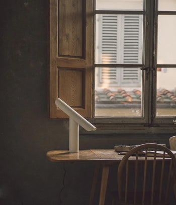 A wooden desk with a FRAMA T-Lamp Table Lamp by FRAMA sits by a window with wooden shutters, overlooking a building with tiled roofing and closed window shutters outside. A wooden chair is tucked beneath the desk.