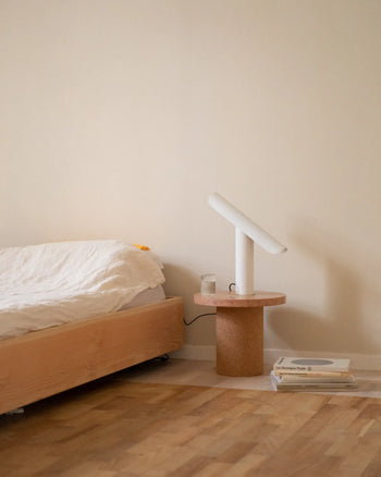 Minimalist bedroom with a wooden bed, white bedding, and a round bedside table featuring the FRAMA T-Lamp Table Lamp by FRAMA. A candle and stacked books rest on the floor, all set against a beige wall and wooden floor.