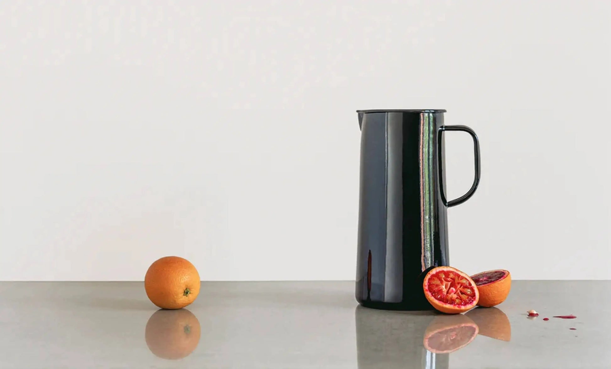 A whole orange, a halved blood orange, and the Falcon Enamelware 3 Pint Jug by Falcon Enamelware are displayed on a reflective gray surface with a plain white background.
