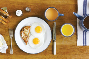 A breakfast plate with two sunny-side-up eggs and buttered toast sits beside a Falcon Enamelware Mug by Falcon Enamelware filled with coffee, plus orange juice, cutlery, and salt and pepper shakers on a wooden table for classic vintage style.