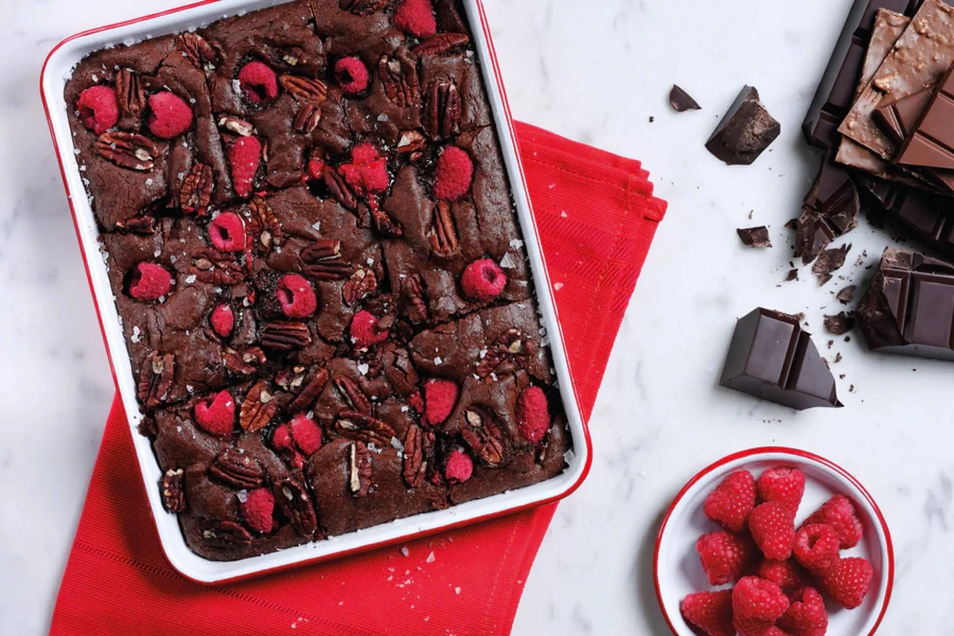 A Falcon Enamelware Serving Tray by Falcon Enamelware holds chocolate brownies topped with raspberries and pecans, set on a red napkin beside a plate of fresh raspberries and chocolate pieces on white marble.