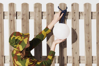 A person in camouflage attire hangs the Fatboy Bolleke, a round white light by Fatboy, with a blue cord on a wooden fence, as sunlight creates modern shadows.