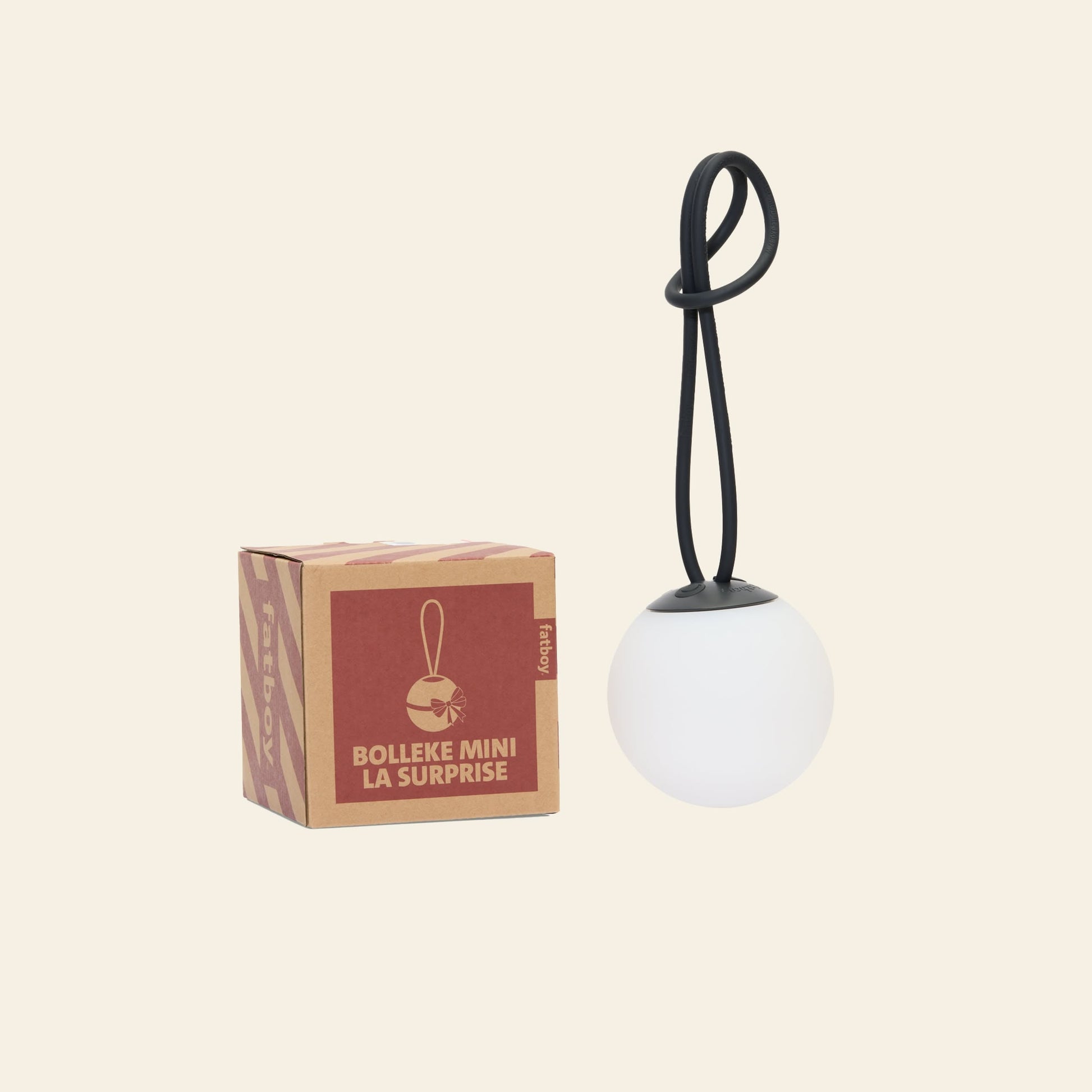 A small Fatboy Bolleke Mini La Surprise box sits beside a round, white Fatboy cordless lamp with a black loop handle, all displayed on a beige background.